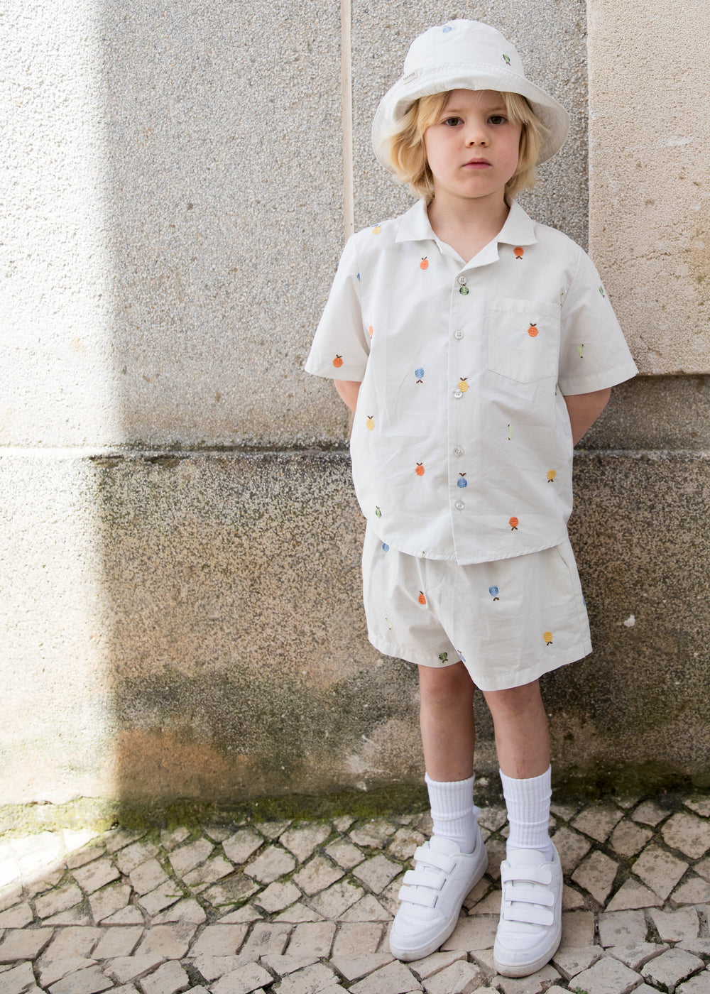 Children wearing a bright set with a shirt and shorts featuring fruit embroidery and a sun hat.