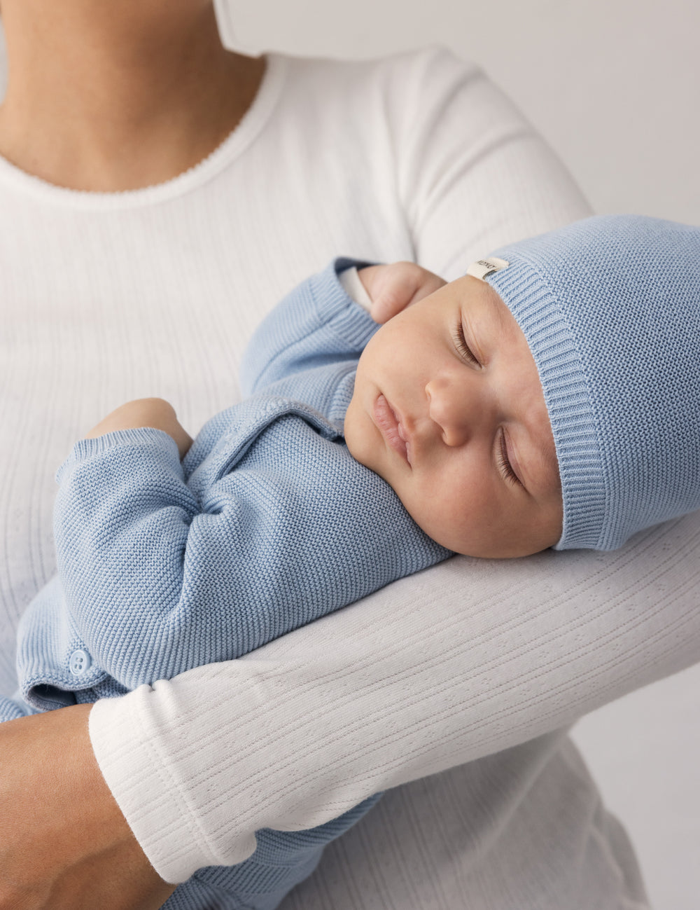 Woman in a white t-shirt holding a sleeping baby wearing a blue hat and a onesie.