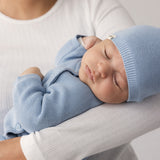 Woman in a white t-shirt holding a sleeping baby wearing a blue hat and a onesie.