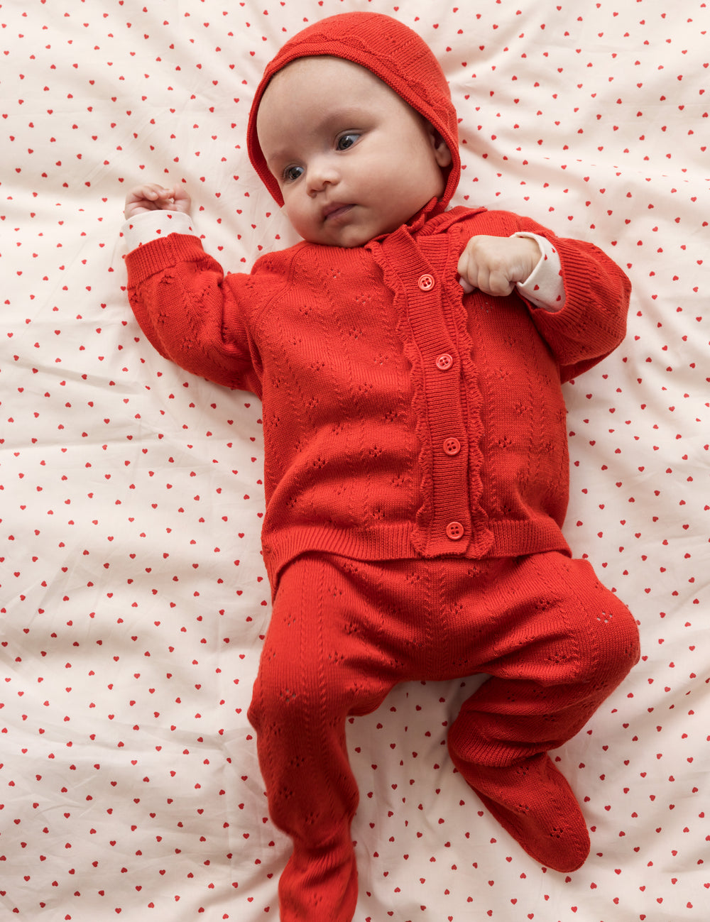 Baby wearing a red cardigan, pants, and matching hat.