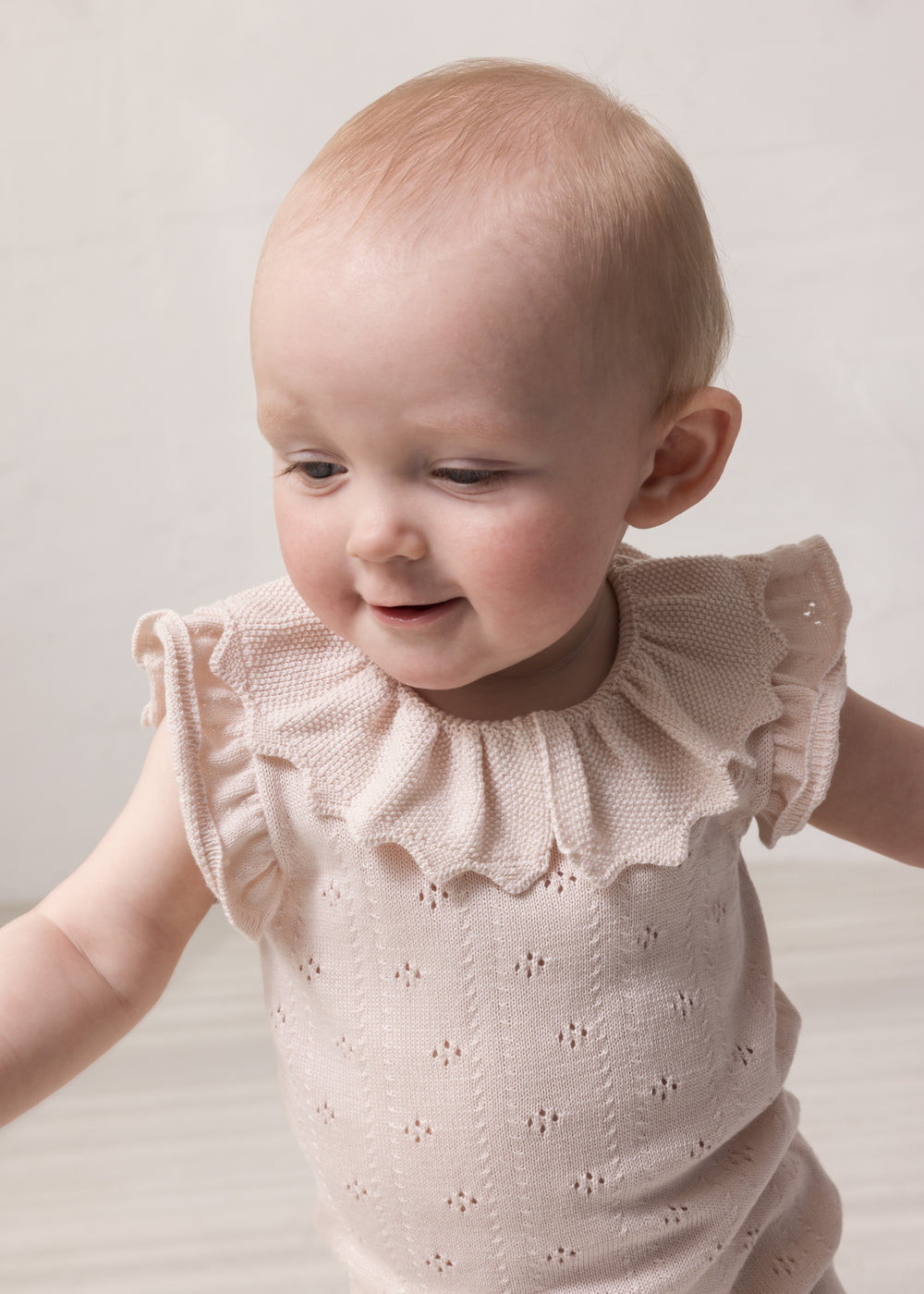 Baby wearing short-sleeved bright red knitted bodystocking with hole pattern and ruffled collar.