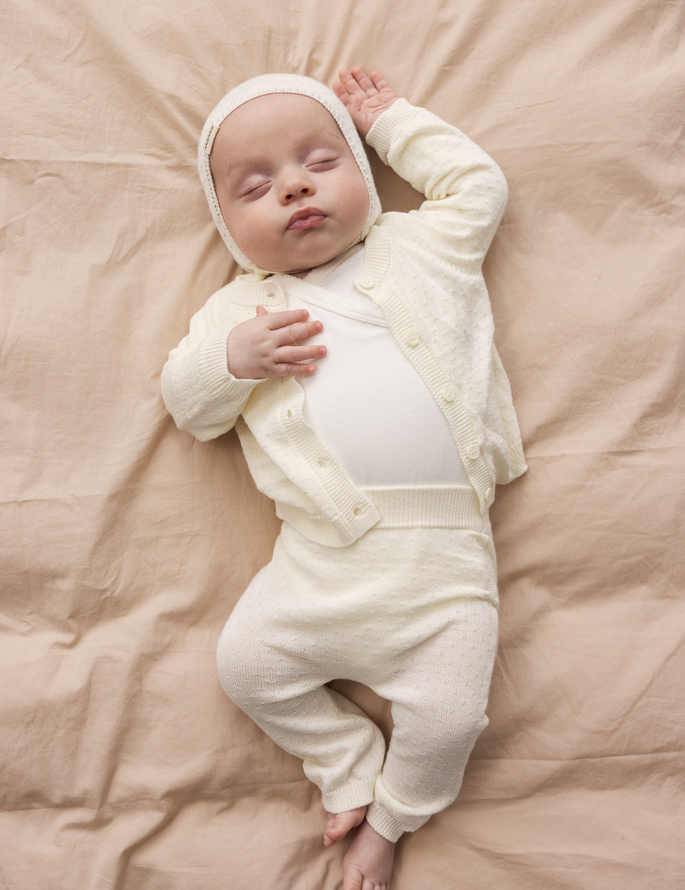 Baby wearing a knitted cardigan with a pretty lace pattern and button closure, matching pants and hat.