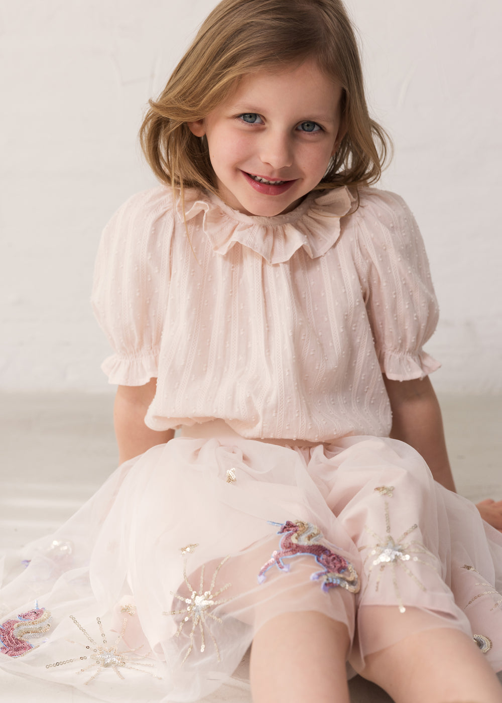 Pige der sidder i en lyserød tyl nederdel  med enhjørninger og stjerner og en lyserød bluse med krave