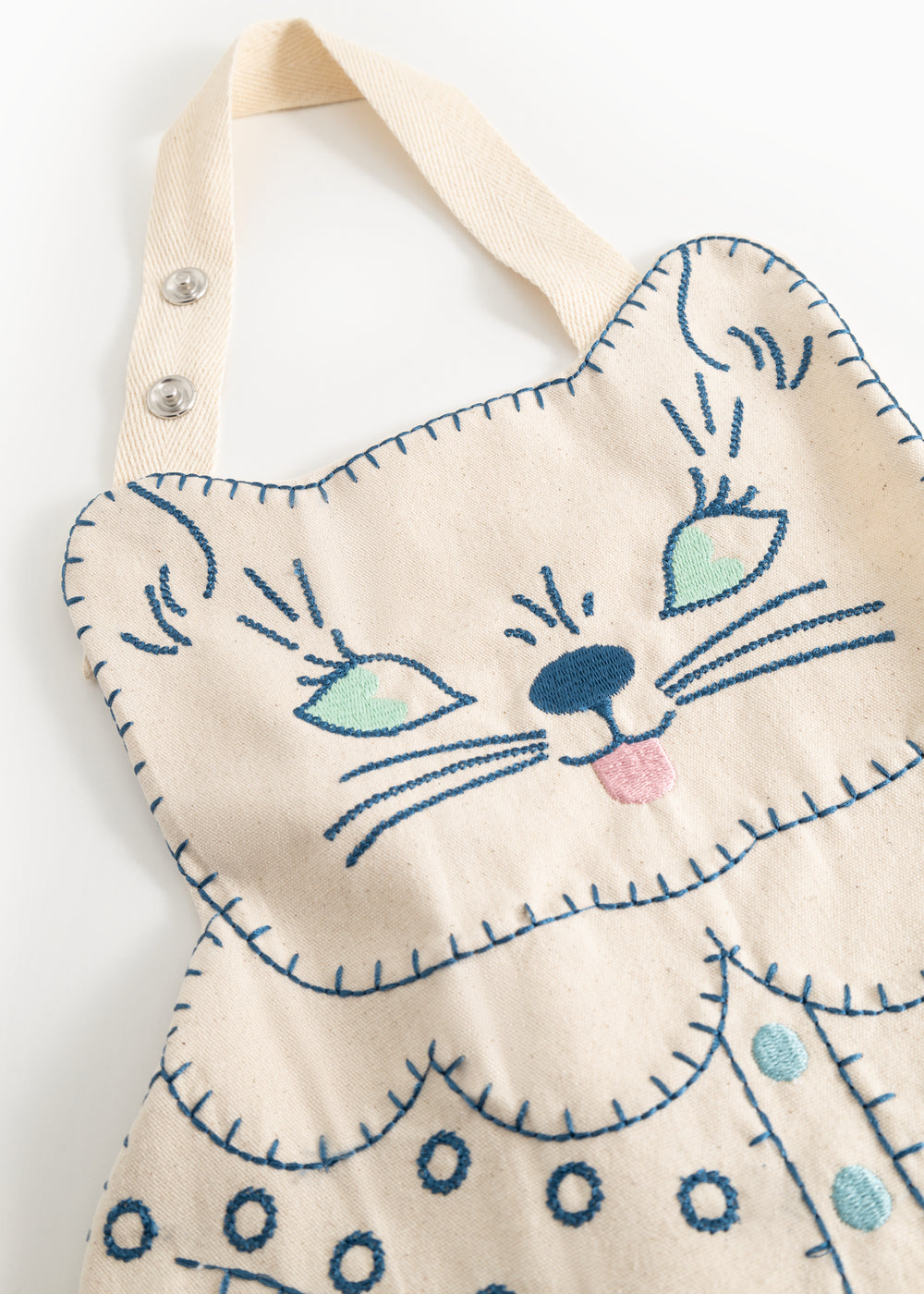 Natural-colored apron with detailed blue cat embroidery and visible stitches along the edges.
