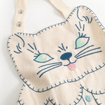 Natural-colored apron with detailed blue cat embroidery and visible stitches along the edges.