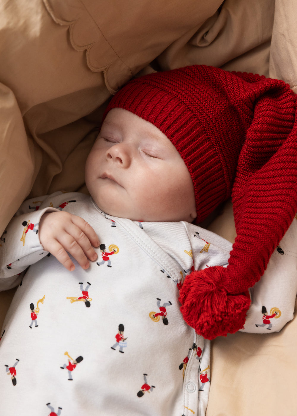 Newborn baby in white jumpsuit with musician print and red hat with tassel.
